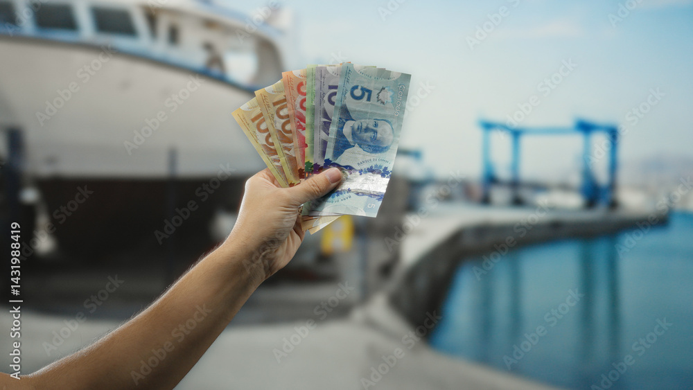 Man holding canadian money outdoors near a seaside port with water and boats, showcasing an economic transaction in a coastal setting.