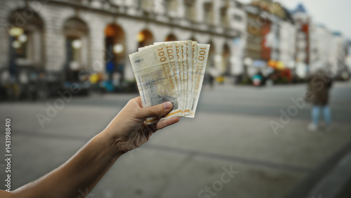 Obraz na płótnie Hand holding danish krone banknotes outdoors in a city street setting, highlighting financial exchange against urban backdrop, with man's figure and historic architecture visible. w salonie