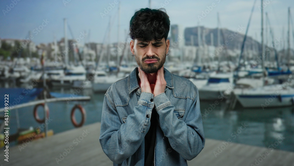 Young man with throat pain stands outdoors near a port with boats and water in the background, demonstrating discomfort against a nautical backdrop.