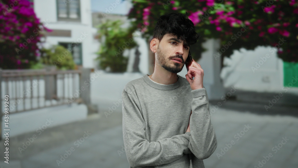 Young man with dark hair and beard in gray sweater talking on smartphone outside on urban street with flowers blurred in background