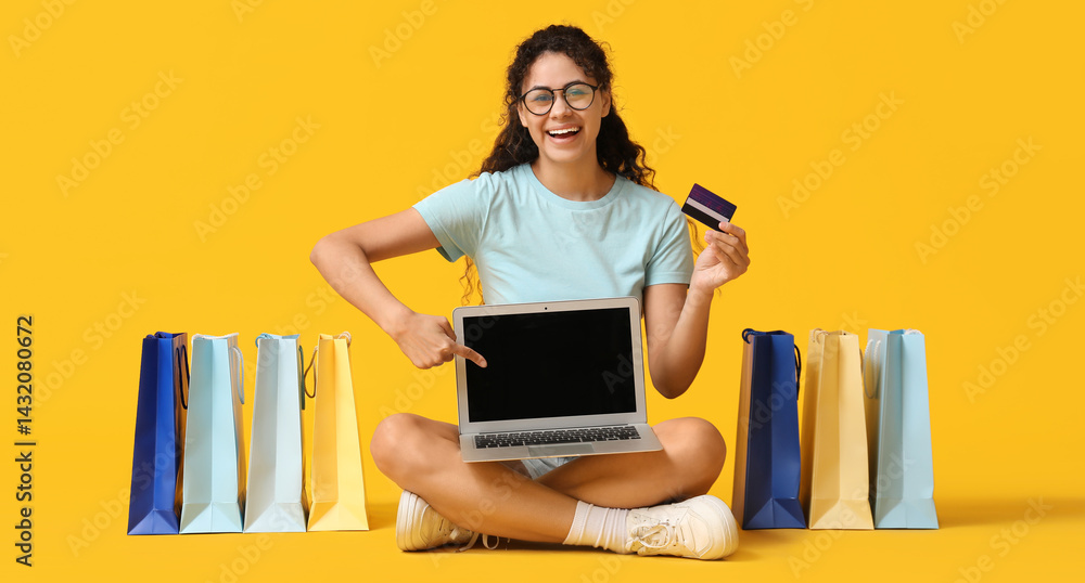 Young woman in eyeglasses sitting with credit cards, laptop and shopping bags on yellow background