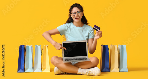 Obraz na płótnie Young woman in eyeglasses sitting with credit cards, laptop and shopping bags on yellow background w salonie