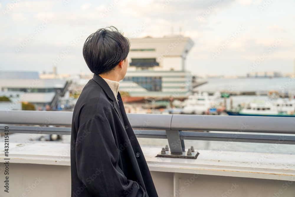 A Japanese man in his twenties wearing a black jacket walks along a seaside path in Minato Mirai, Yokohama, Kanagawa Prefecture, enjoying the breeze, city buildings, and the calm harbor atmosphere.