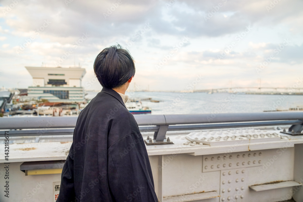 A Japanese man in his twenties wearing a black jacket walks along a seaside path in Minato Mirai, Yokohama, Kanagawa Prefecture, enjoying the breeze, city buildings, and the calm harbor atmosphere.