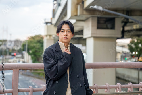 Obraz na płótnie A Japanese man in his twenties wearing a black jacket is in Minato Mirai, Yokohama, Kanagawa Prefecture. He sits on a bench, gazing into the distance, surrounded by the calm mood of the modern city. w salonie