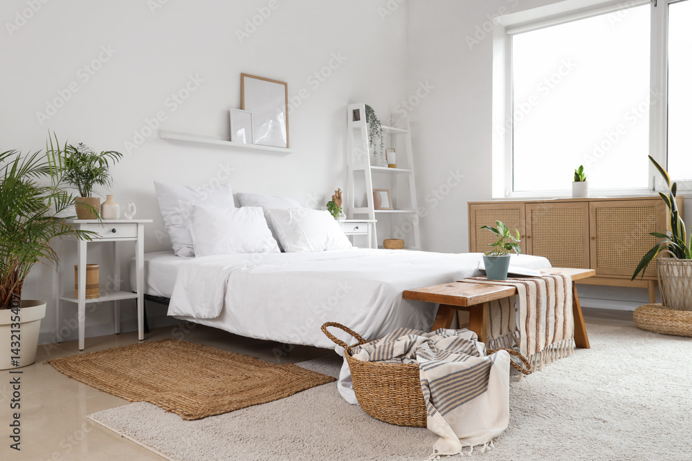 Obraz premium Stylish interior of light bedroom with wooden chest of drawers, bench, houseplants and bed