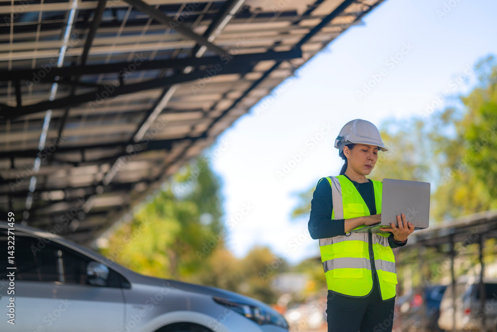 custom made wallpaper toronto digitalA female technician wearing a safety helmet and reflective vest uses a laptop while inspecting a solar carport installation. The scene features solar panels and parked cars under a clear sky.