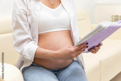 Obraz na płótnie Pregnant woman sitting on sofa in clinic, holding medical record and waiting for doctor's appointment. Happy young expectant mother. Family planning and IVF centers concept. High quality photo w salonie