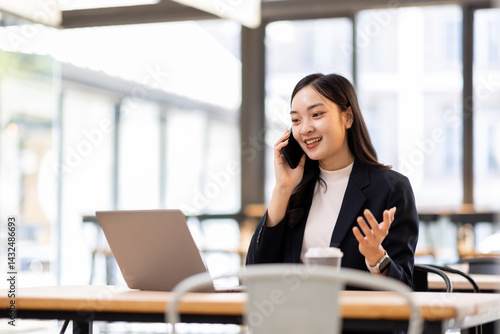 Happy young business woman manager talking consulting client on cell phone working on laptop in office. Busy woman professional business making call on cell using computer sitting at desk
