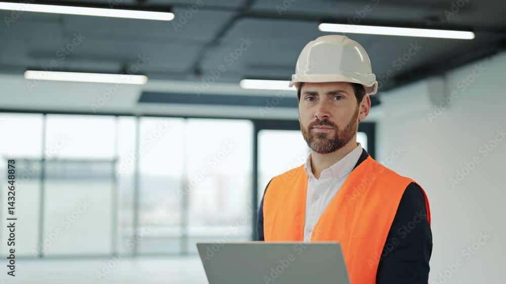 Successful engineer in an orange vest and a white helmet works on a laptop in an empty office building. He smiles with satisfaction. Successful engineer in an orange vest and a white helmet works on a laptop in an empty office building. He smiles with satisfaction.