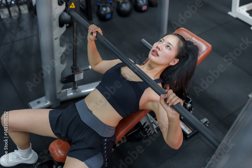 Young woman exercising on a bench press machine, lifting a weighted bar to strengthen her upper body muscles, showcasing dedication to fitness and healthy lifestyle in a modern gym setting