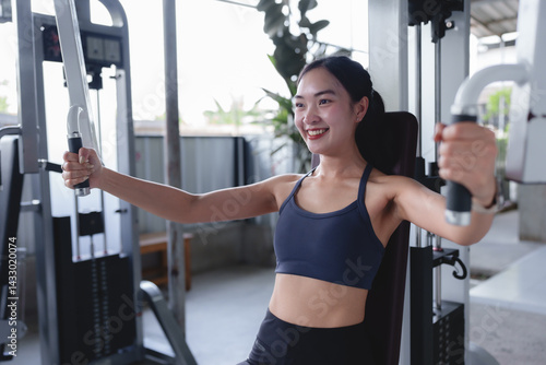 Smiling asian fitness model performing a chest fly exercise on a pec deck machine, focusing on building upper body strength and muscle definition in a modern gym environment