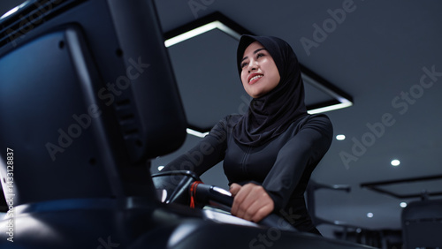A smiling Southeast Asian Muslim woman in a black sports hijab works out on a treadmill in a modern gym. The photo reflects positivity, health, and the joy of movement in an inclusive fitness 