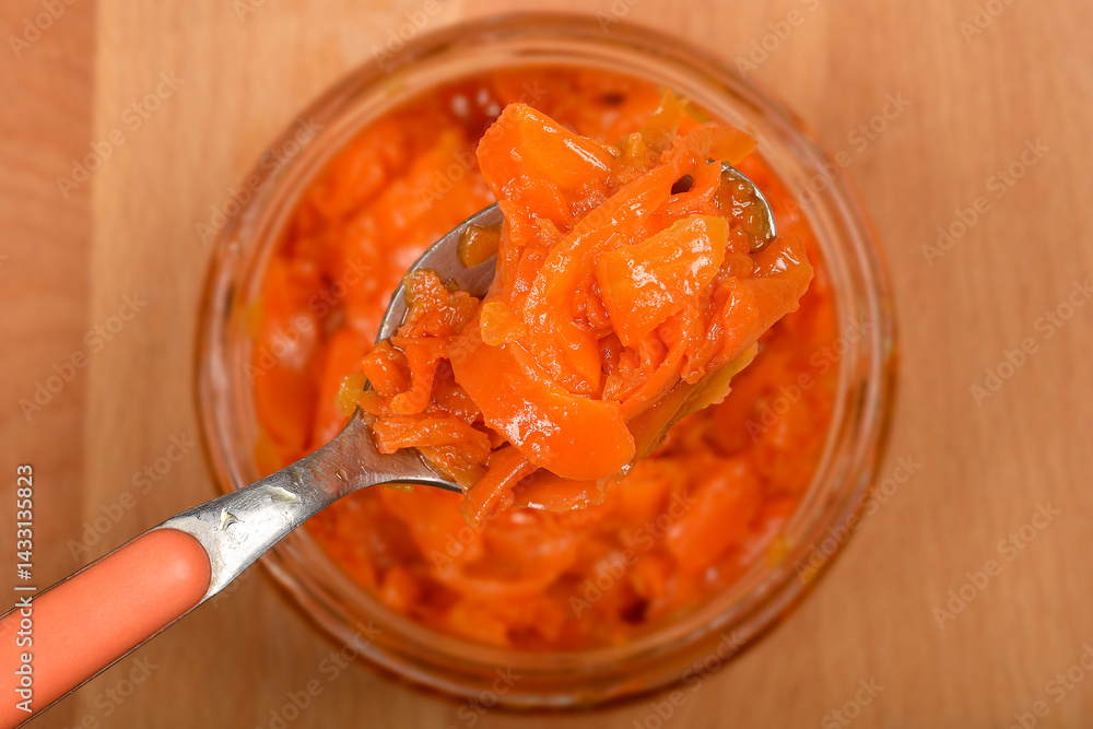 A spoonful of vibrant orange, thinly sliced carrots prepared as vegan salmon is lifted from a clear glass jar against a light brown wooden background - mrkva pripremljena kao veganski losos