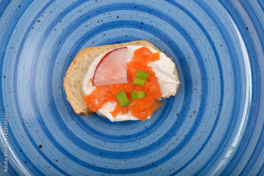 An overhead shot shows a canape with creamy white spread, vibrant orange carrot prepared as vegan salmon, a slice of radish, and green herbs on a blue patterned plate