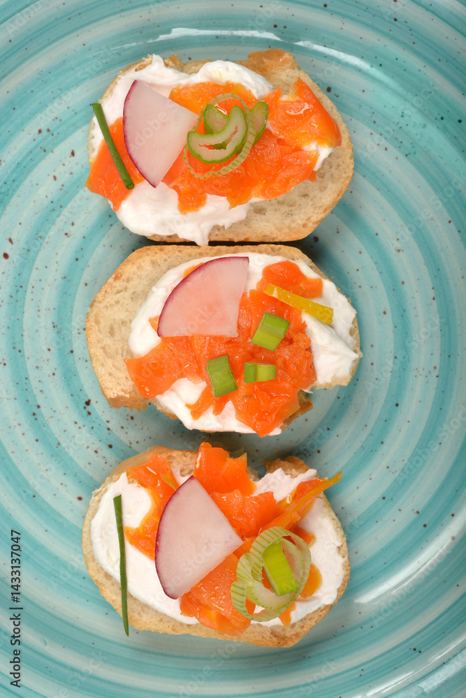 An overhead view shows three elegant canapes with a creamy spread, vibrant orange carrot prepared as vegan salmon, radish slices, and green chives on a light blue swirled plate