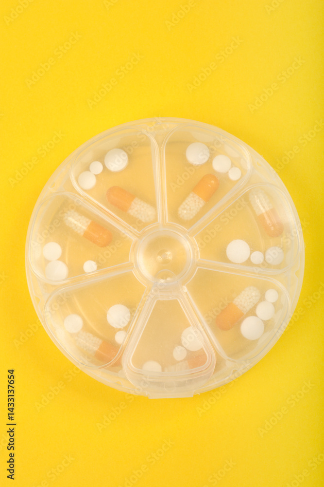 A clear, round pill organizer with seven compartments holds various white and orange capsules and tablets against a bright yellow background