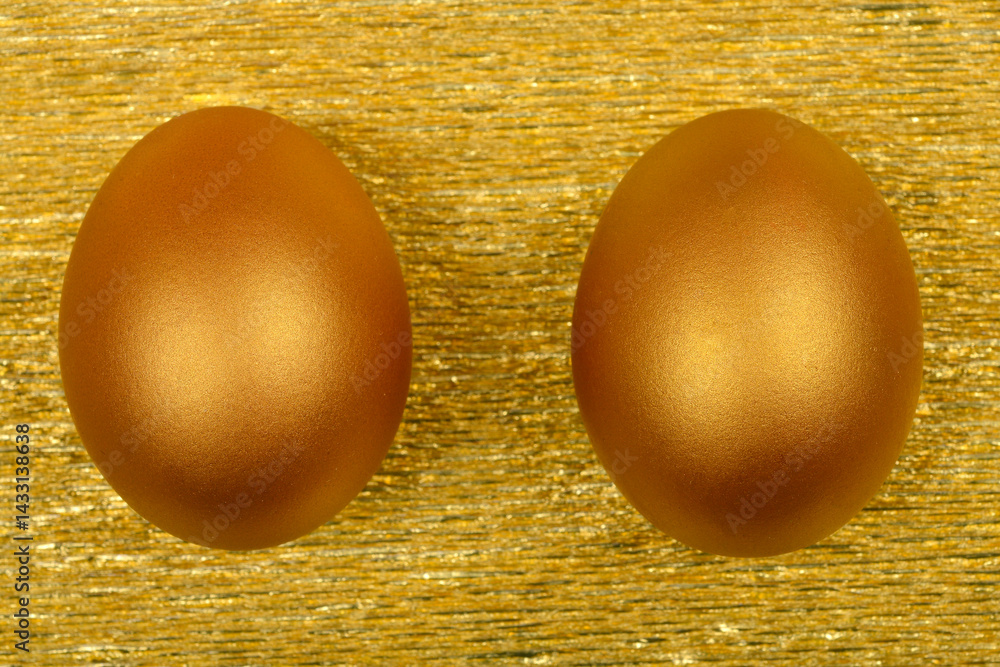Two metallic golden eggs are positioned side-by-side on a textured, shimmering gold surface