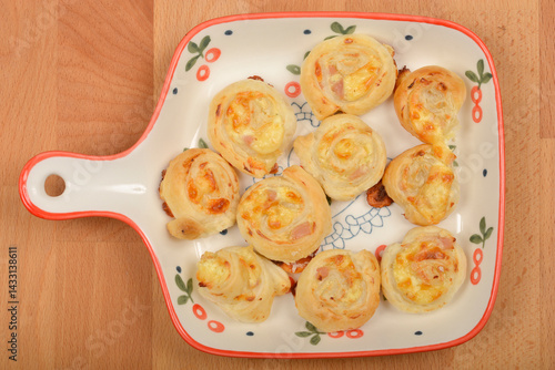 Golden-brown puff pastry rolls filled with cheese and ham are arranged on a decorative, handled plate atop a wooden surface
