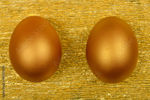 Two metallic golden eggs are positioned side-by-side on a textured, shimmering gold surface