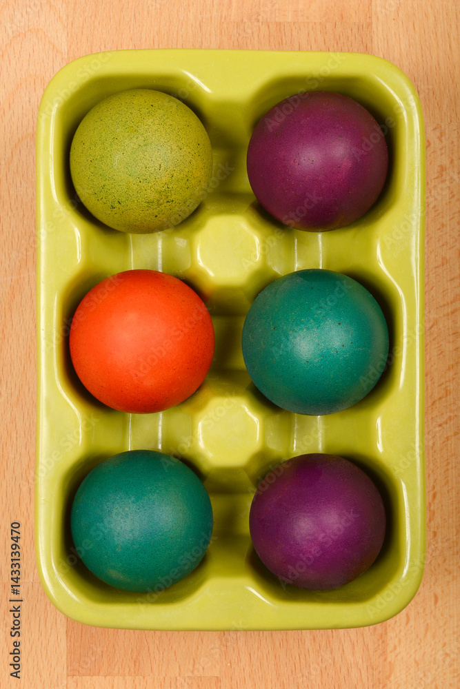 A top-down view shows a light green ceramic egg carton holding six colorful Easter eggs on a light brown wooden surface