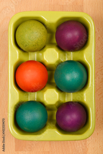 A top-down view shows a light green ceramic egg carton holding six colorful Easter eggs on a light brown wooden surface