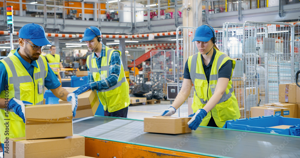 In a Modern Sorting Center Facility, Diverse Workers in Safety Vests Loading Parcels Onto a Conveyor, Efficiently Working Together as a Team. Logistics and Postal Service Operations In a Modern Sorting Center Facility, Diverse Workers in Safety Vests Loading Parcels Onto a Conveyor, Efficiently Working Together as a Team. Logistics and Postal Service Operations