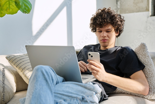 Handsome young man with laptop using smartphone sitting on sofa at home