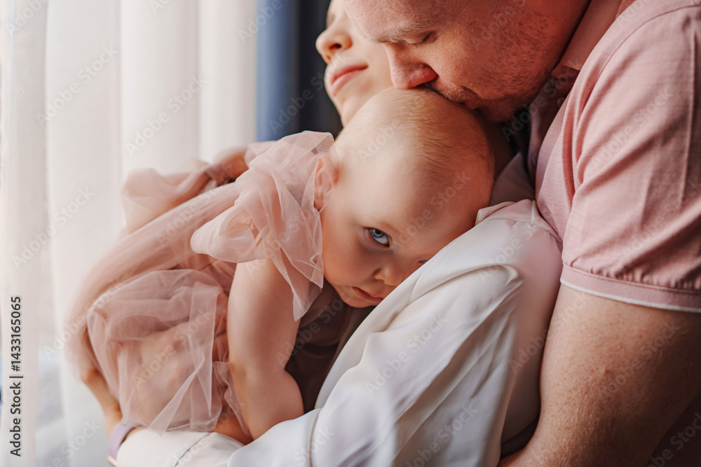 Happy and gentle parents hold baby daughter in pink dress in their arms.