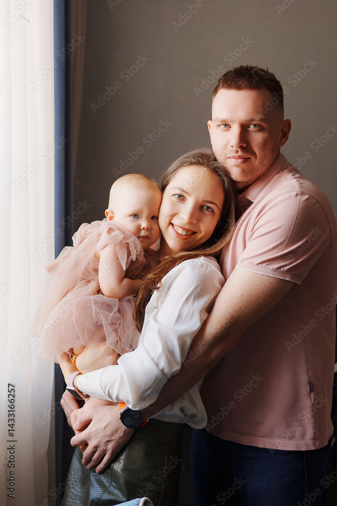 Happy and gentle parents hold baby daughter in pink dress in their arms.