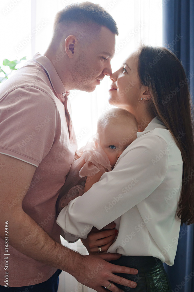 Happy and gentle parents hold baby daughter in pink dress in their arms.