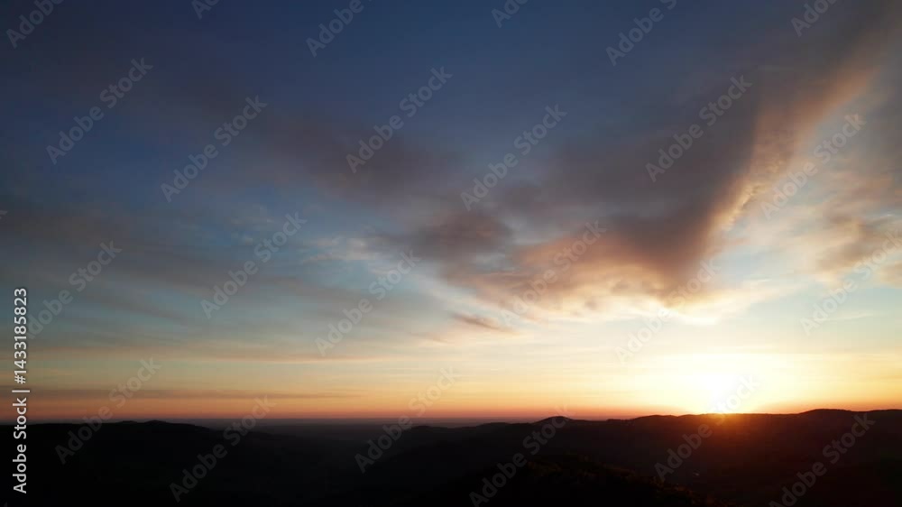 Sun sets behind rolling hills, casting warm, golden glow across sky. Wispy clouds, tinged with hues of orange and yellow, stretch across deepening blue sky. Serene and expansive landscape. Sun sets behind rolling hills, casting warm, golden glow across sky. Wispy clouds, tinged with hues of orange and yellow, stretch across deepening blue sky. Serene and expansive landscape.