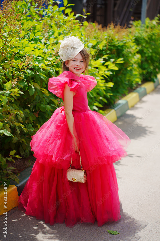 A girl in a lush pink dress, hat and with a golden handbag near the green bushes