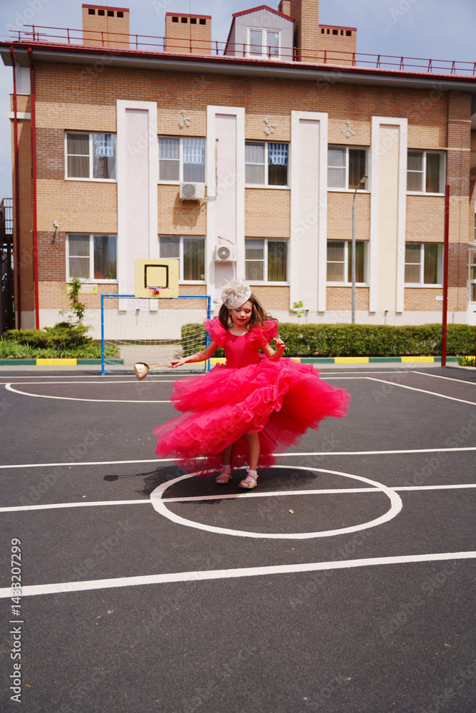 A cute little girl in an elegant lush pink dress is playing on the sports ground