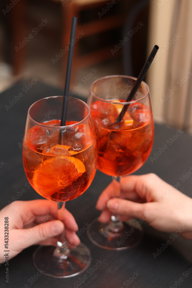Two glasses with Aperol Spritz in the hands of girls. Cold refreshing alcoholic cocktail Aperol Spritz. Cocktails close-up with selective focus. Alcoholic drink with a straw. Traditional Italian drink