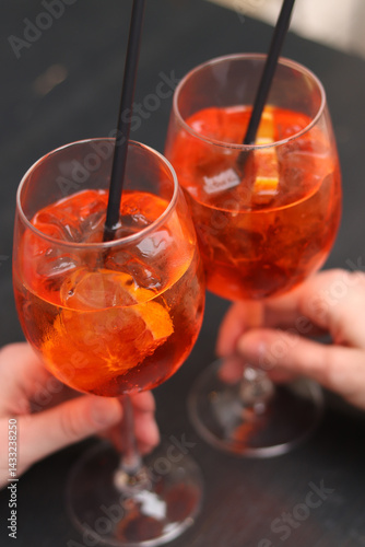 Two glasses with Aperol Spritz in the hands of girls. Cold refreshing alcoholic cocktail Aperol Spritz. Cocktails close-up with selective focus. Alcoholic drink with a straw. Traditional Italian drink
