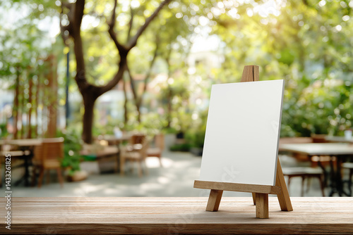 Blank white sign on small wooden easel in outdoor cafe - Empty mockup template with natural green bokeh background and wooden tabletop