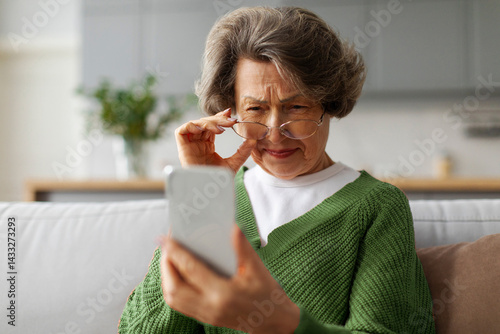 Elderly European woman squinting eyes trying read message on cellphone screen, sitting on couch at home. Bad vision problem