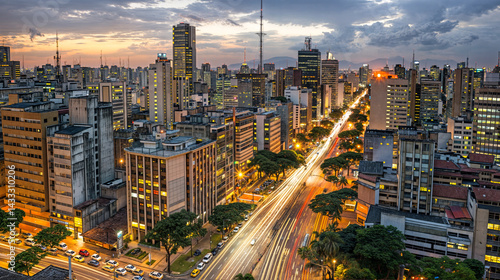 A bustling cityscape at dusk, with tall buildings illuminated by street lights and car headlights creating streaks of light on the roads. 