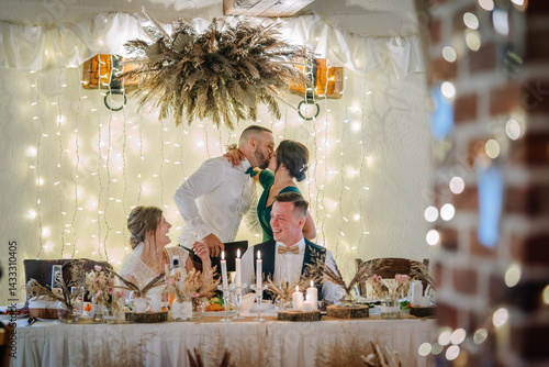 Φωτογραφία Joyful wedding moment with guests cheering as a couple kisses under romantic fairy lights, seated at a rustic, candlelit reception table