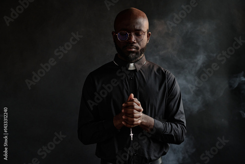 Medium shot of young African American priest in black cassock holding rosary with cross while he praying silently. He in dark room with incense hovering in air