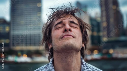 Young man with messy hair and closed eyes smiling deeply while standing outdoors enjoying peace and fresh air in downtown city street.