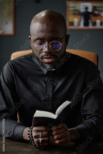 Vertical shot of African American priest wearing round glasses and black cassock sitting in comfortable office chair and reading bible