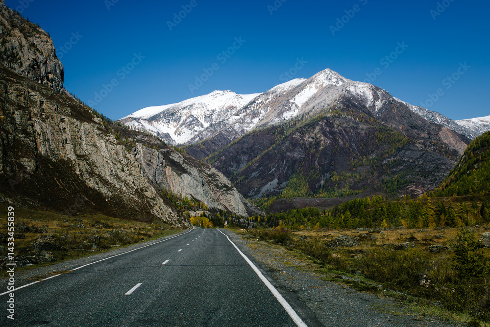 A mountain road winding through the rugged terrain of the Altai Republic, surrounded by harsh, snow-covered peaks and dramatic natural beauty.