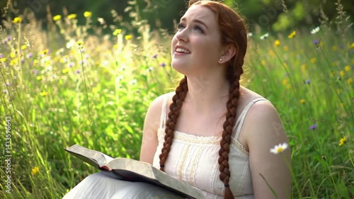 Young christian woman reading a bible book on a meadow of wildflowers in a park outdoors, looking up to heaven with gratitude to God