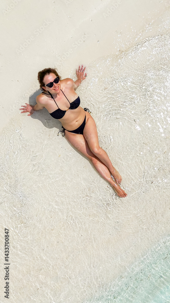 Fototapeta premium woman in black swimsuit lies on wet sand as gentle waves touch the shore, girl is sunbathing on white sand, enjoying the sun, tropical background, aerial view, beachwear, summer travel, relaxation