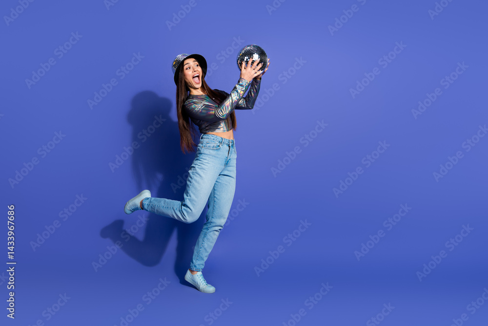 Obraz premium Young woman posing with a fun attitude holding a shiny disco ball against a vibrant purple background wearing trendy casual fashion.