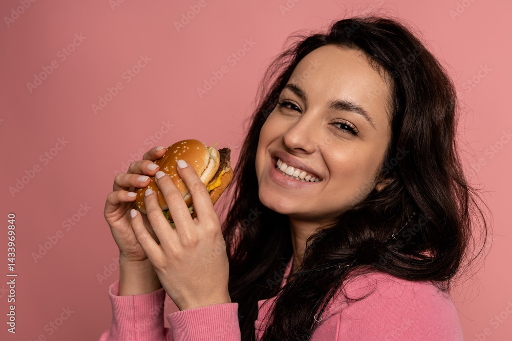 Pleased brunette with a delicious cheeseburger in her hands smiling during the studio photo shoot. Appetite and favorite fast food concept Pleased brunette with a delicious cheeseburger in her hands smiling during the studio photo shoot. Appetite and favorite fast food concept
