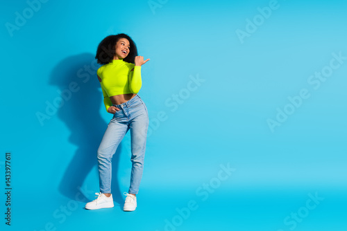 Smiling young woman in neon green top and casual jeans posing confidently against a vibrant blue background with gesture