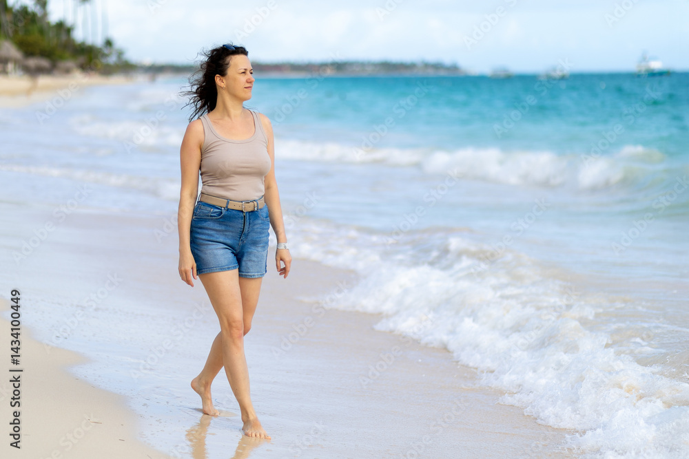 Woman walking barefoot along a serene beach during a sunny day with gentle waves in the background Woman walking barefoot along a serene beach during a sunny day with gentle waves in the background